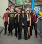 Preview: Foto der Band Arte Bella & The Fellas. Eine Gruppe von acht Personen steht auf einem urbanen Gehweg mit Graffiti-Wänden im Hintergrund. Alle Personen schauen freundlich in die Kamera. Die Gruppe ist gemischt, mit Männern und Frauen, alle in lockerer, indi